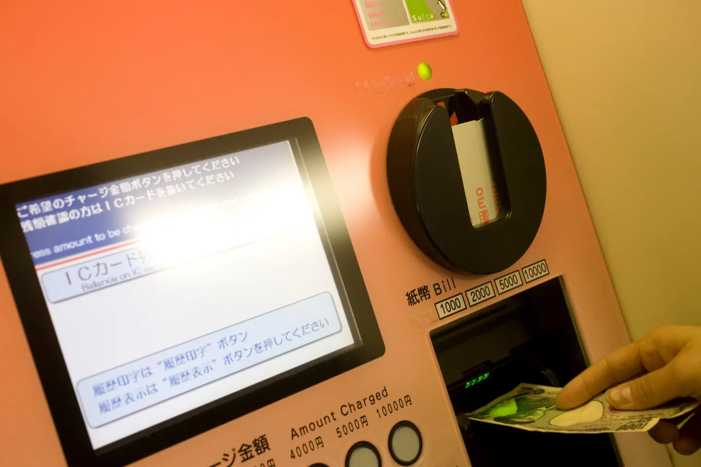 يد تُدخل ورقة 1,000 ين في ماكينة شحن بطاقة Suica داخل محطة يابانية